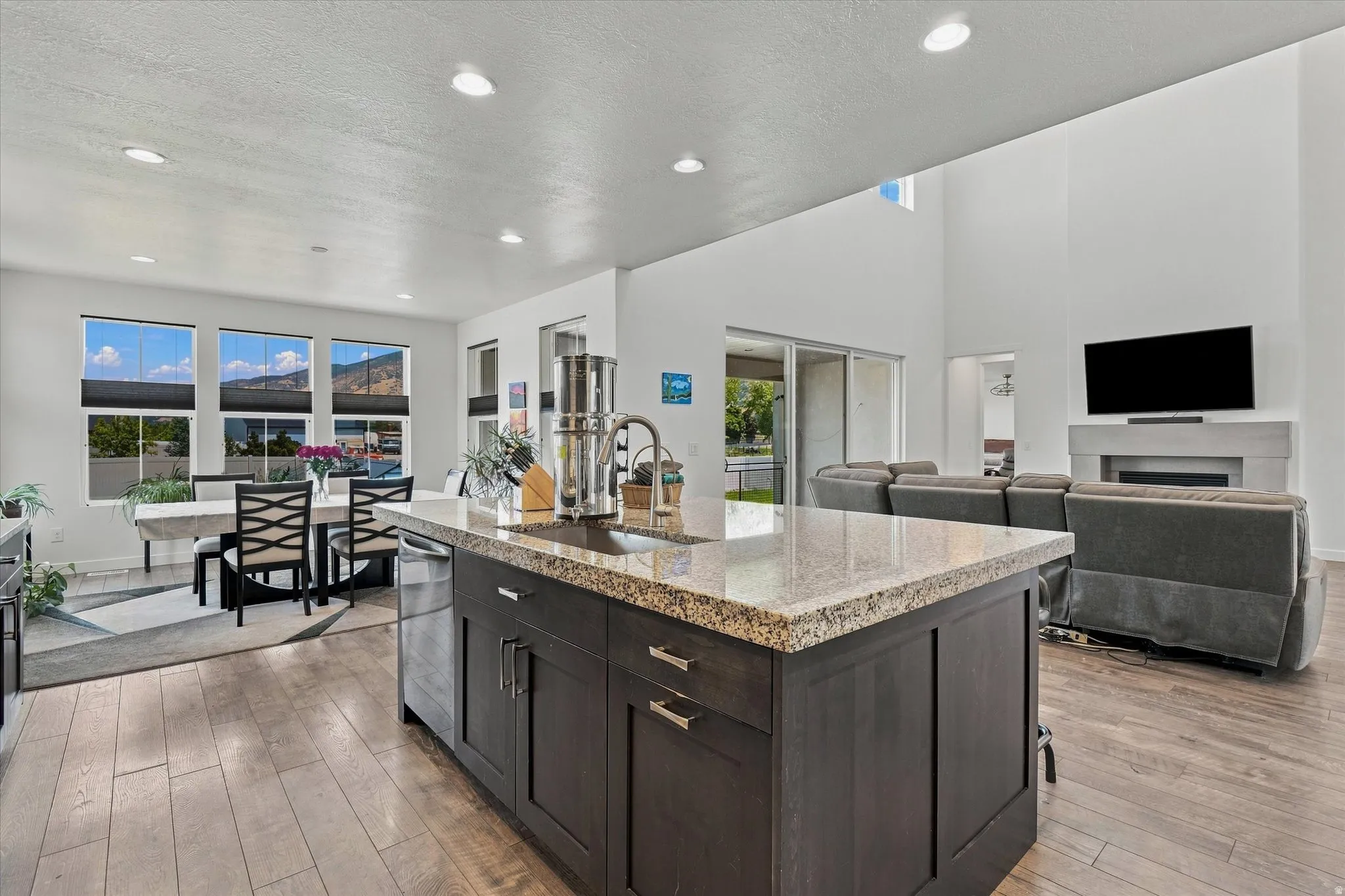 Spacious open-concept living area featuring an eat-in kitchen island with a granite countertop, wood-finish flooring, recessed lighting, and a two-story living room with a fireplace