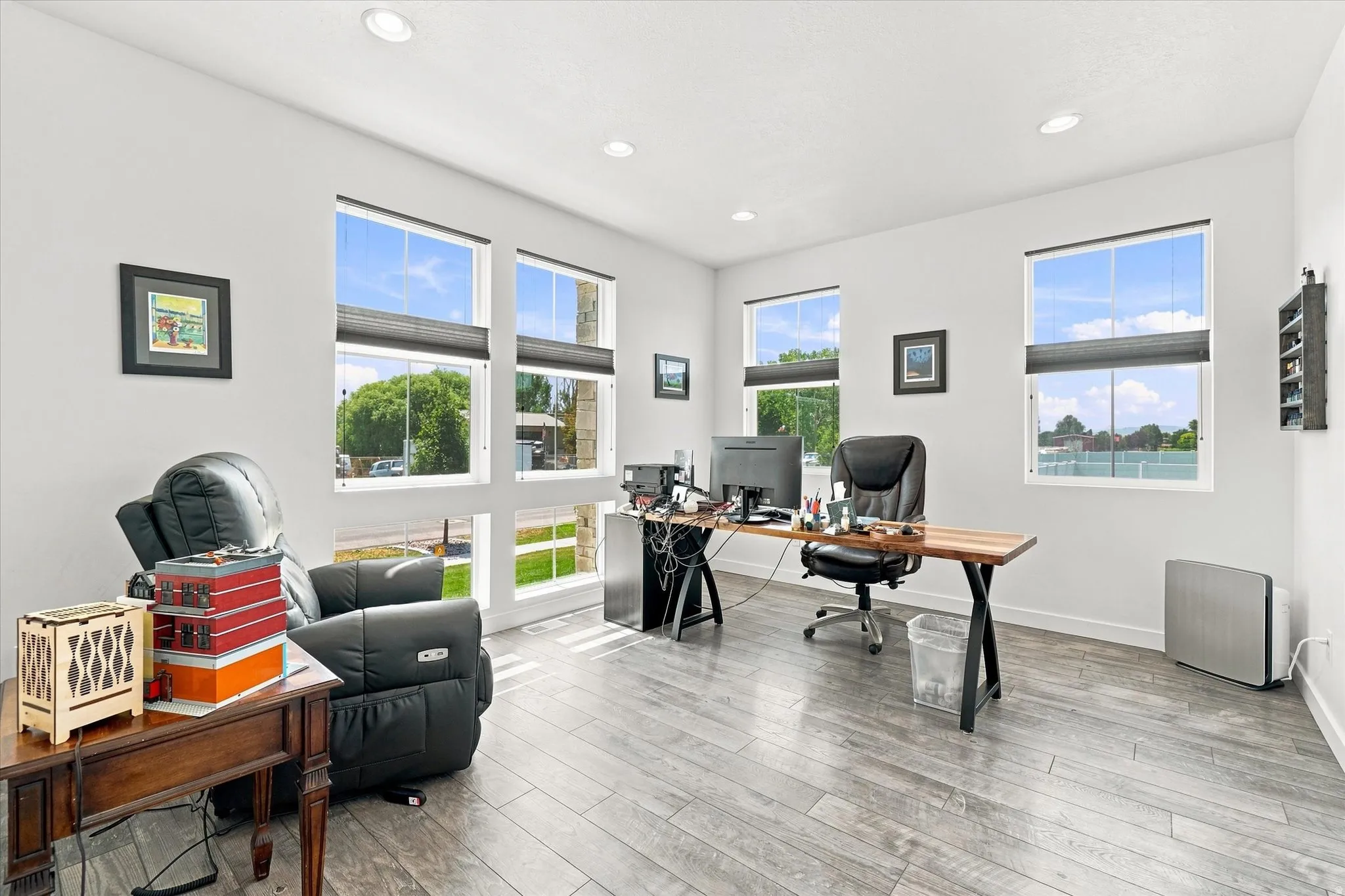 Bright, light-filled home office featuring expansive multi-pane windows with natural light, wood-finish flooring, recessed ceiling lighting, and a spacious desk workspace