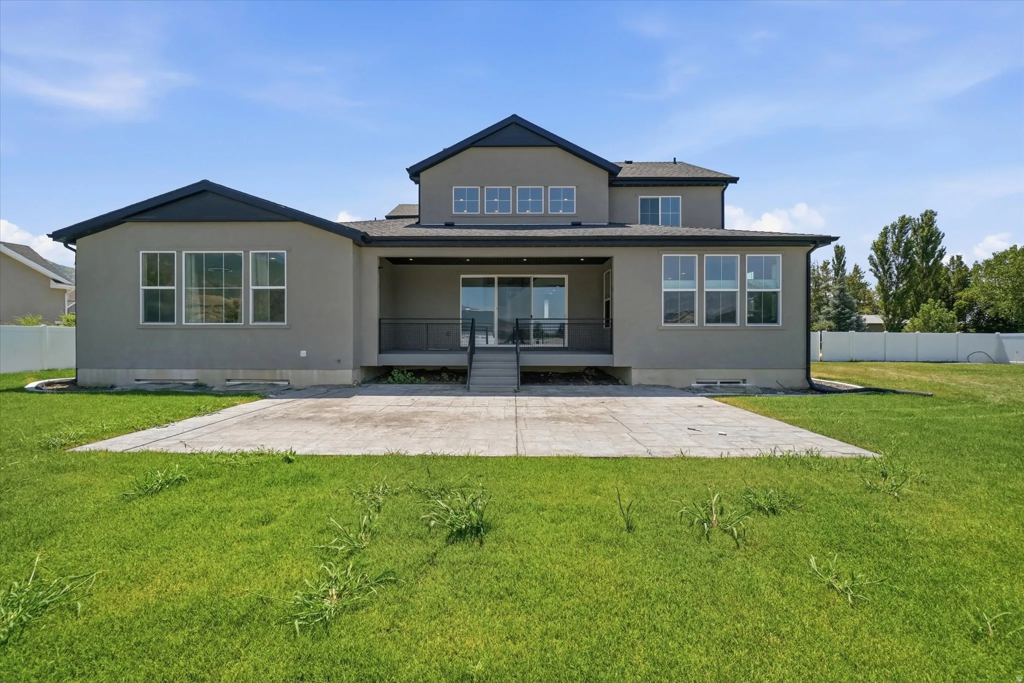 Rear exterior featuring a large concrete patio, covered deck with sliding glass doors, multiple large windows, and a multi-level roofline