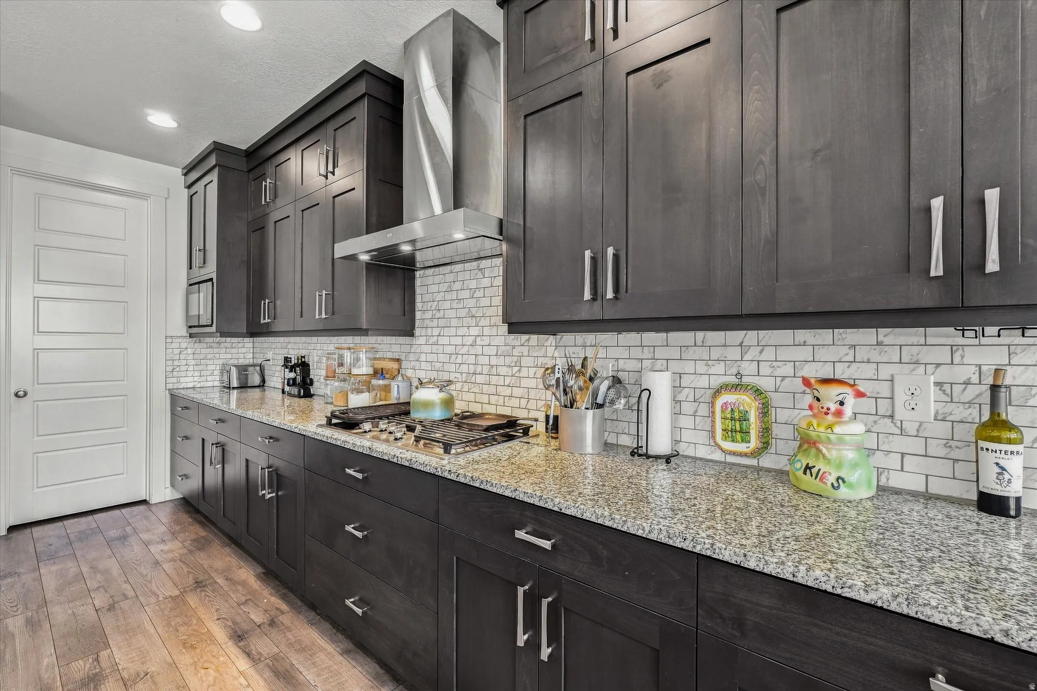 Modern kitchen featuring dark wood-finish cabinetry, light speckled countertops, a white subway tile backsplash, and wood-finish flooring