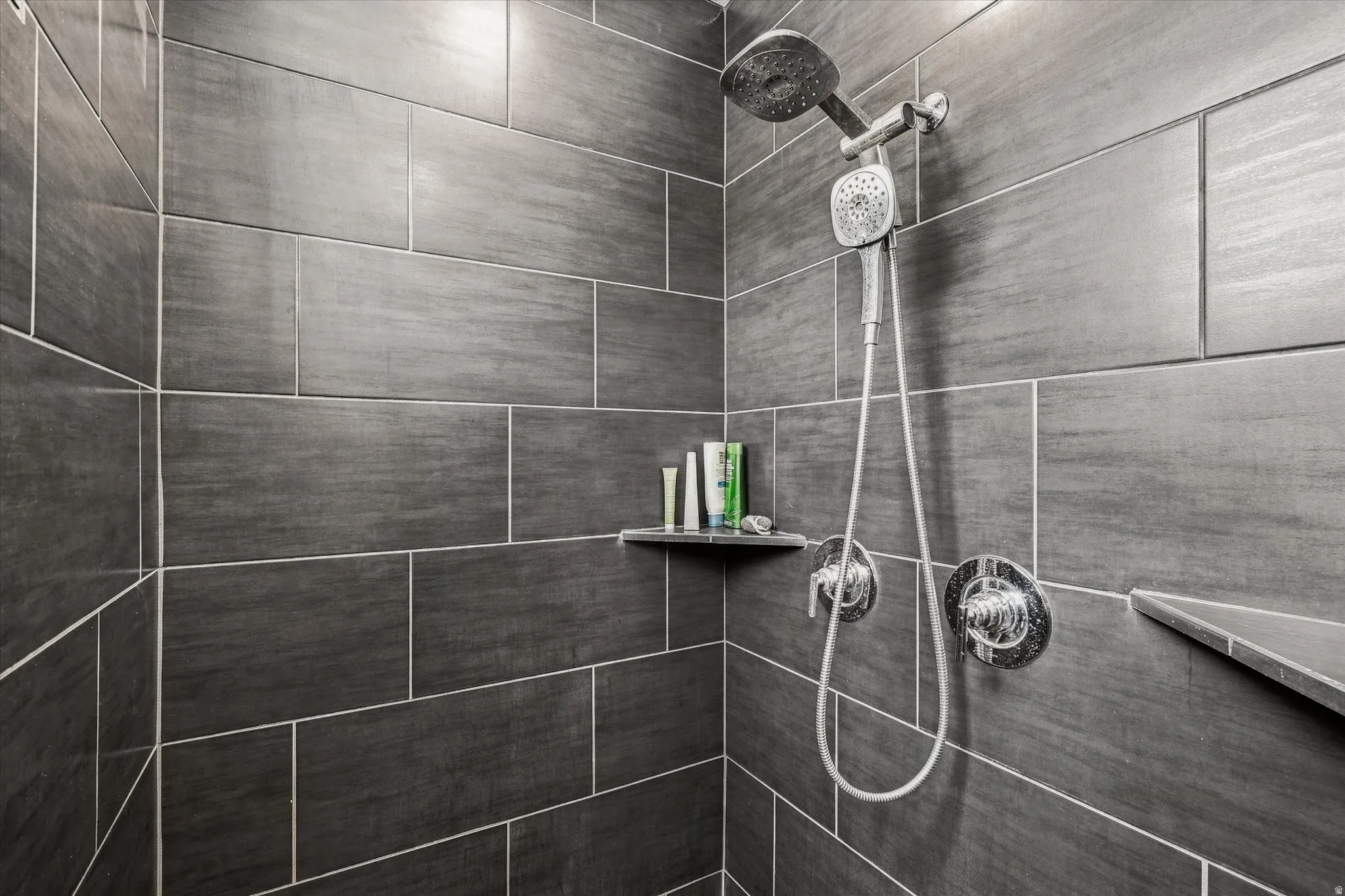 Shower enclosure featuring dark gray rectangular tile with light grout, a rainfall showerhead, a handheld showerhead, and integrated shelving