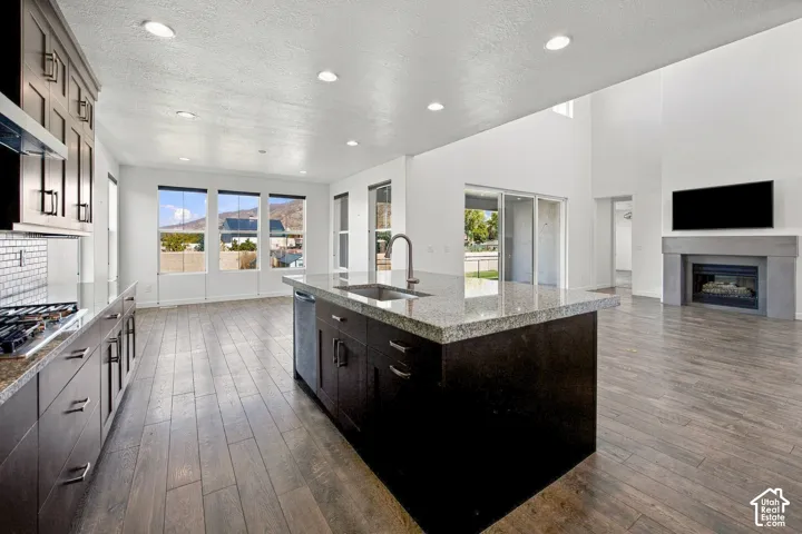 Open-concept kitchen and living area featuring a large center island with dark cabinetry and stone countertop, stainless steel appliances, white subway tile backsplash, wide-plank engineered wood flooring, recessed lighting throughout, and a gas fireplace