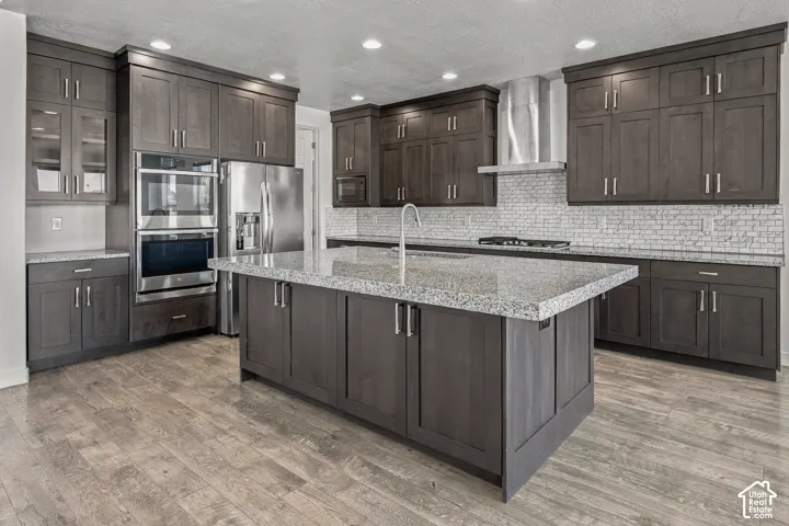 Kitchen featuring engineered wood flooring, dark wood cabinetry, a central island with extra thick granite countertop, stainless steel appliances, and a tiled backsplash