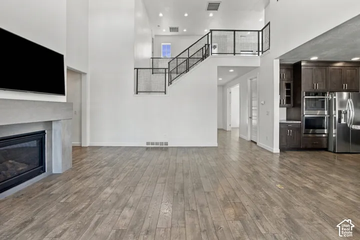 Enginnered wood flooring, a contemporary fireplace with a concrete mantel, a modern staircase with black metal railing, a kitchen area with dark wood cabinetry, and stainless steel appliances