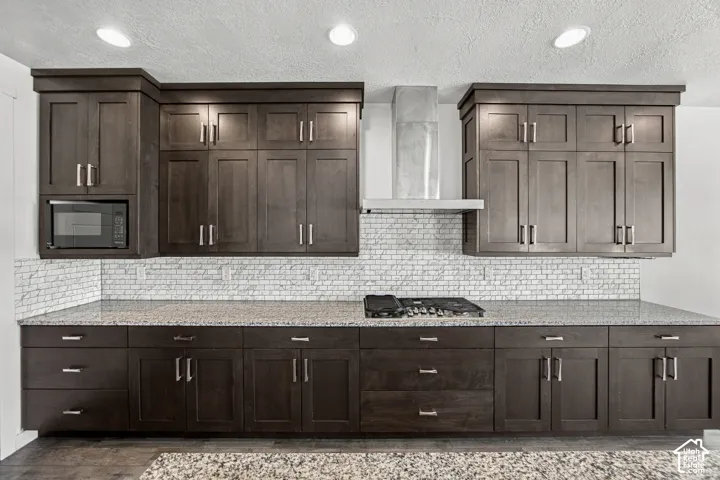 Dark wood-finish cabinets galore, light speckled countertops, subway tile backsplash, stainless steel microwave, and a stainless steel range hood