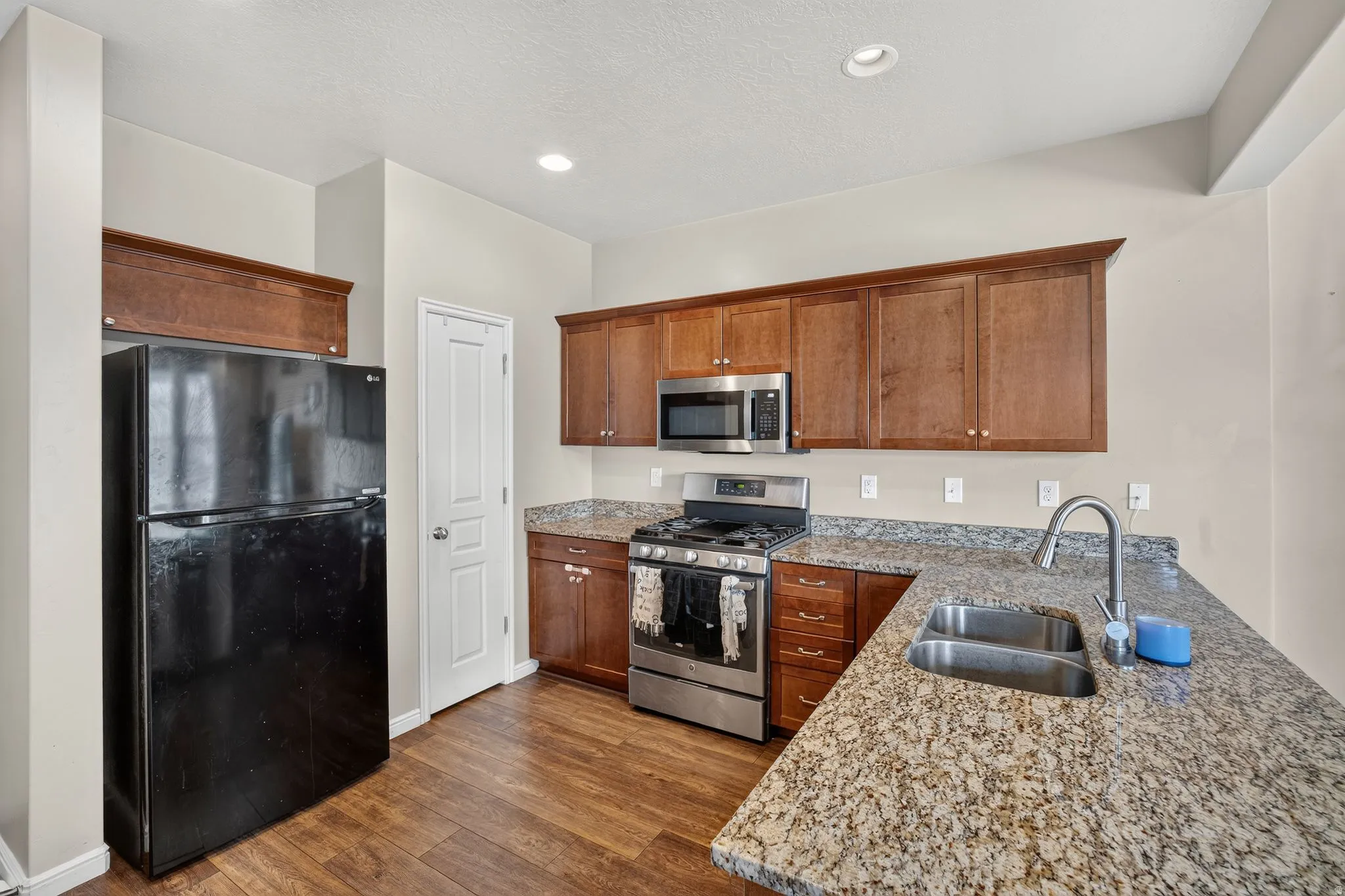 Kitchen featuring wood-finish flooring, L-shaped speckled countertops, a double-basin stainless steel sink, and wood cabinetry with stainless steel appliances