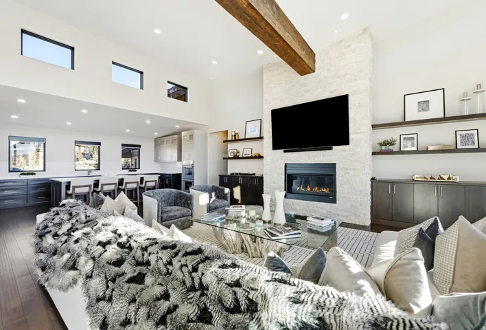 Spacious living area featuring a stone-clad fireplace with built-in shelving, high ceilings, exposed wood beam, and dark wood-finish flooring