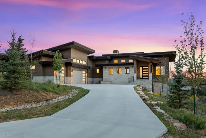 Modern architectural exterior featuring a concrete driveway, multi-paneled garage doors, a contemporary entry door, stonework accents, and varied natural landscaping