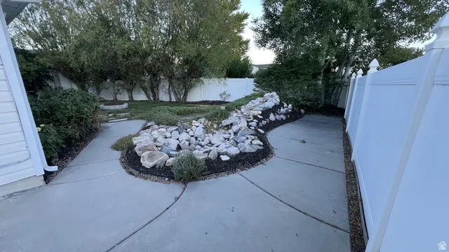 Landscaped backyard featuring a winding concrete pathway, decorative rock garden with dark mulch, mature trees, and a white privacy fence