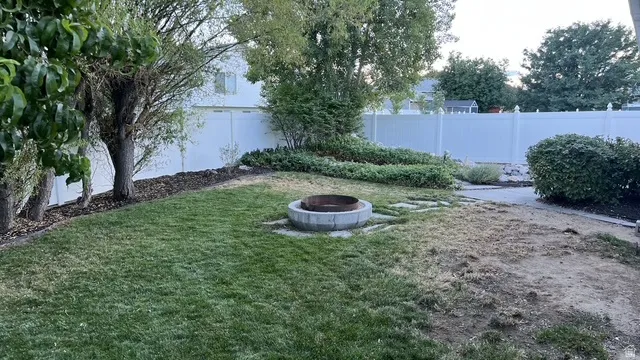 Lawn area featuring a built-in stone fire pit, surrounded by mature trees, privacy fencing, and garden beds