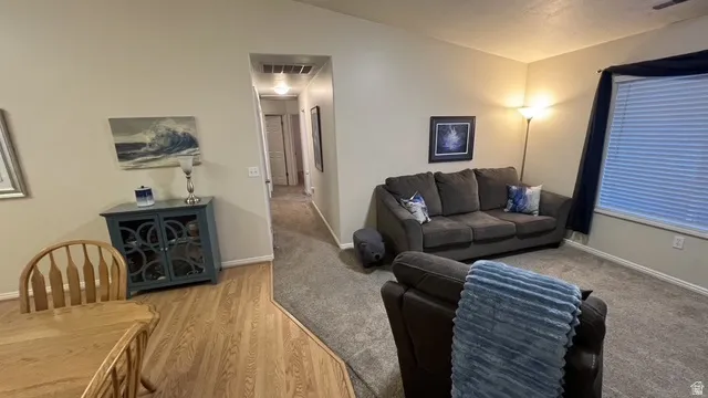 Open-concept living area featuring wood-finish flooring, a carpeted lounge space, a window with blinds, and a hallway entry