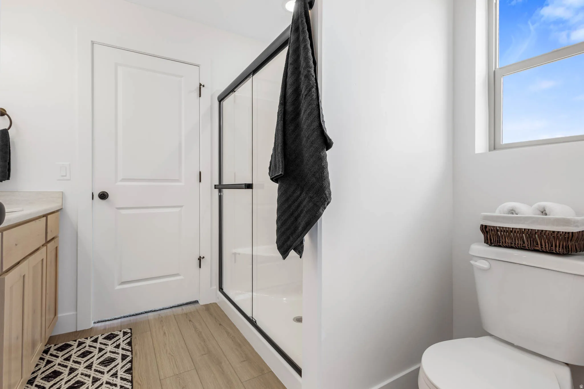 Bathroom featuring a walk-in shower with a black-framed glass enclosure, light wood-finish flooring, a single window, and a light wood vanity with a light-toned countertop