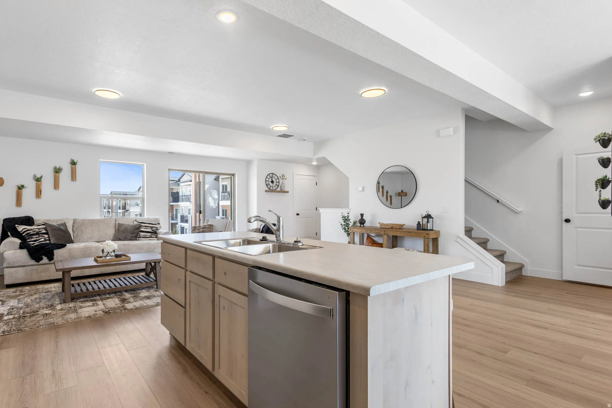 Bright open-concept living space featuring wood-finish flooring, recessed lighting, a kitchen island with integrated sink, and an adjacent living area with a sliding glass door to an outdoor space