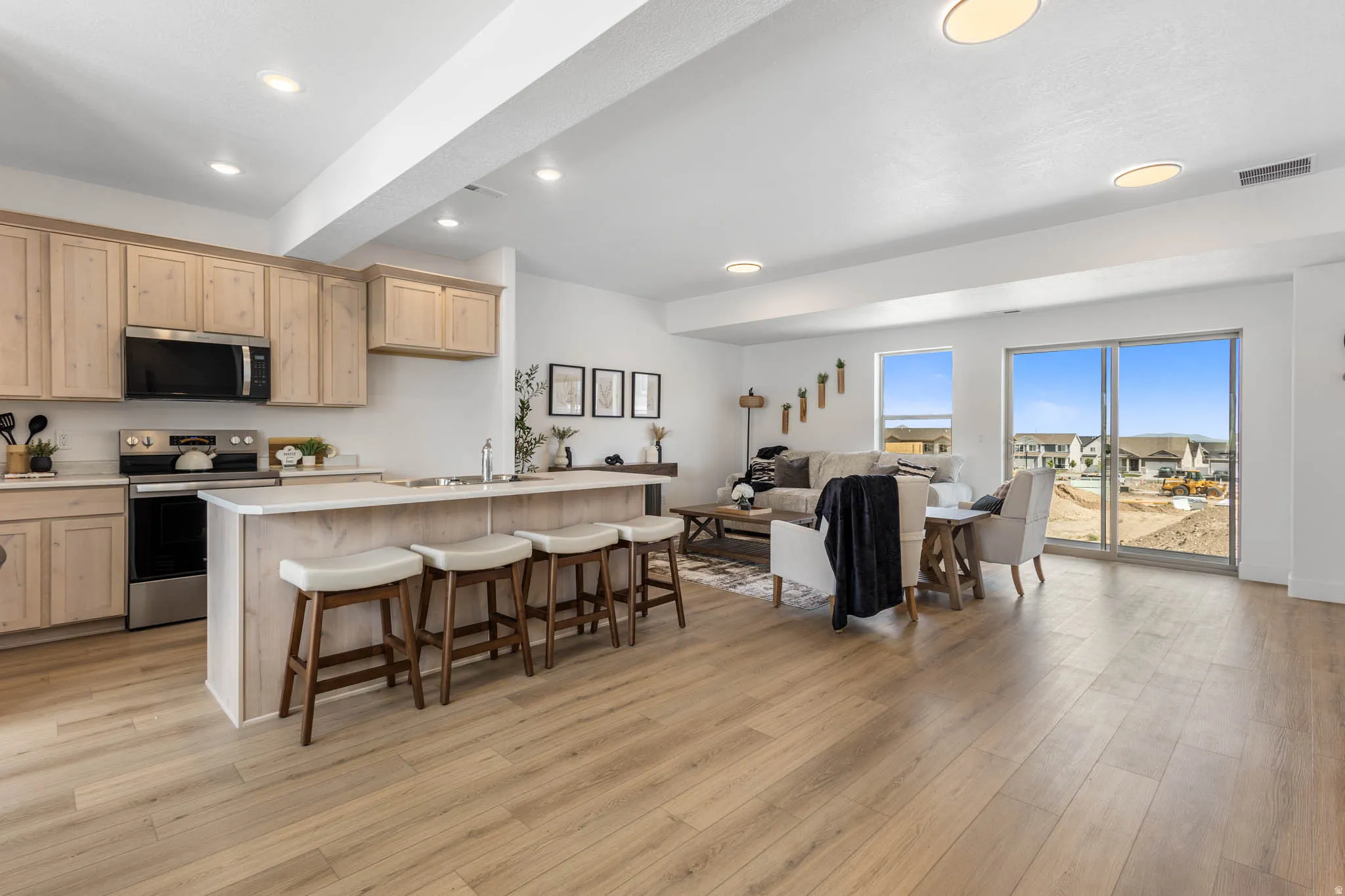 Open-concept living area featuring light wood-finish flooring, a kitchen island with seating, natural wood cabinetry, stainless steel appliances, and abundant natural light from large windows and sliding glass doors