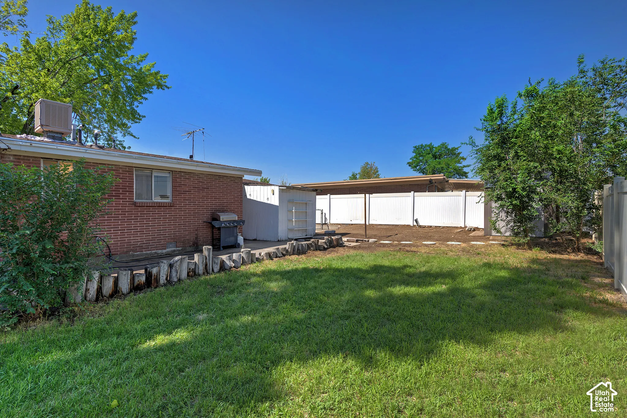 Expansive green lawn area with a partial brick exterior, a white privacy fence, a storage shed, and mature trees