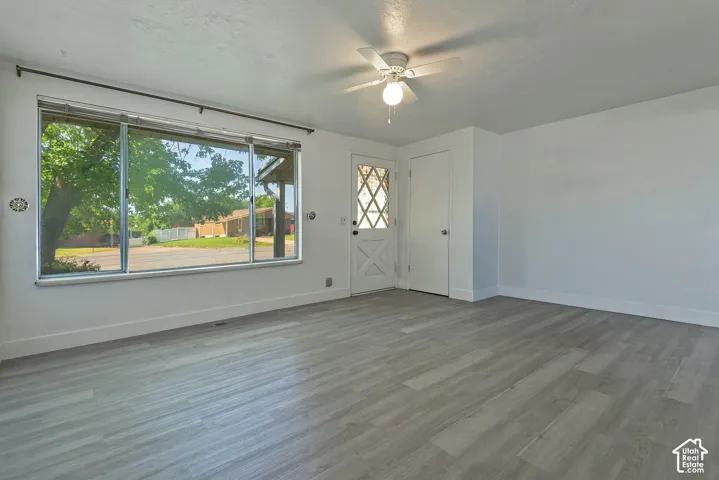 Spacious room featuring light wood-finish flooring, a large picture window, a white front door with decorative glass, a flush-mounted ceiling fan with lighting, and crisp white walls