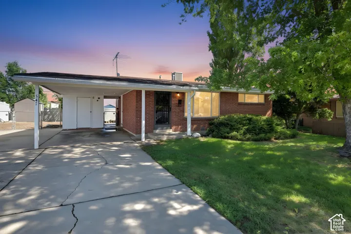 Brick ranch-style home featuring an attached carport, covered entry, front yard landscaping, and mature trees