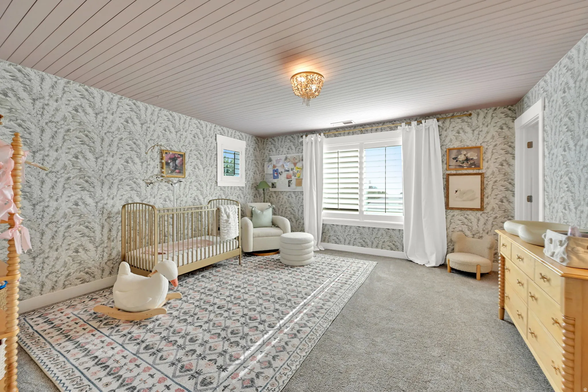 Spacious room featuring patterned wallpaper, textured ceiling, a window with white louvered shutters, light gray carpeting, and a decorative chandelier