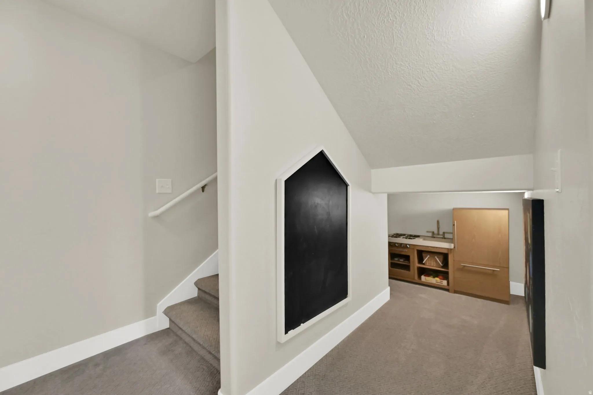 Carpeted hallway with a house-shaped chalkboard accent wall, leading to a kitchenette featuring a built-in refrigerator, open shelving, and a sink