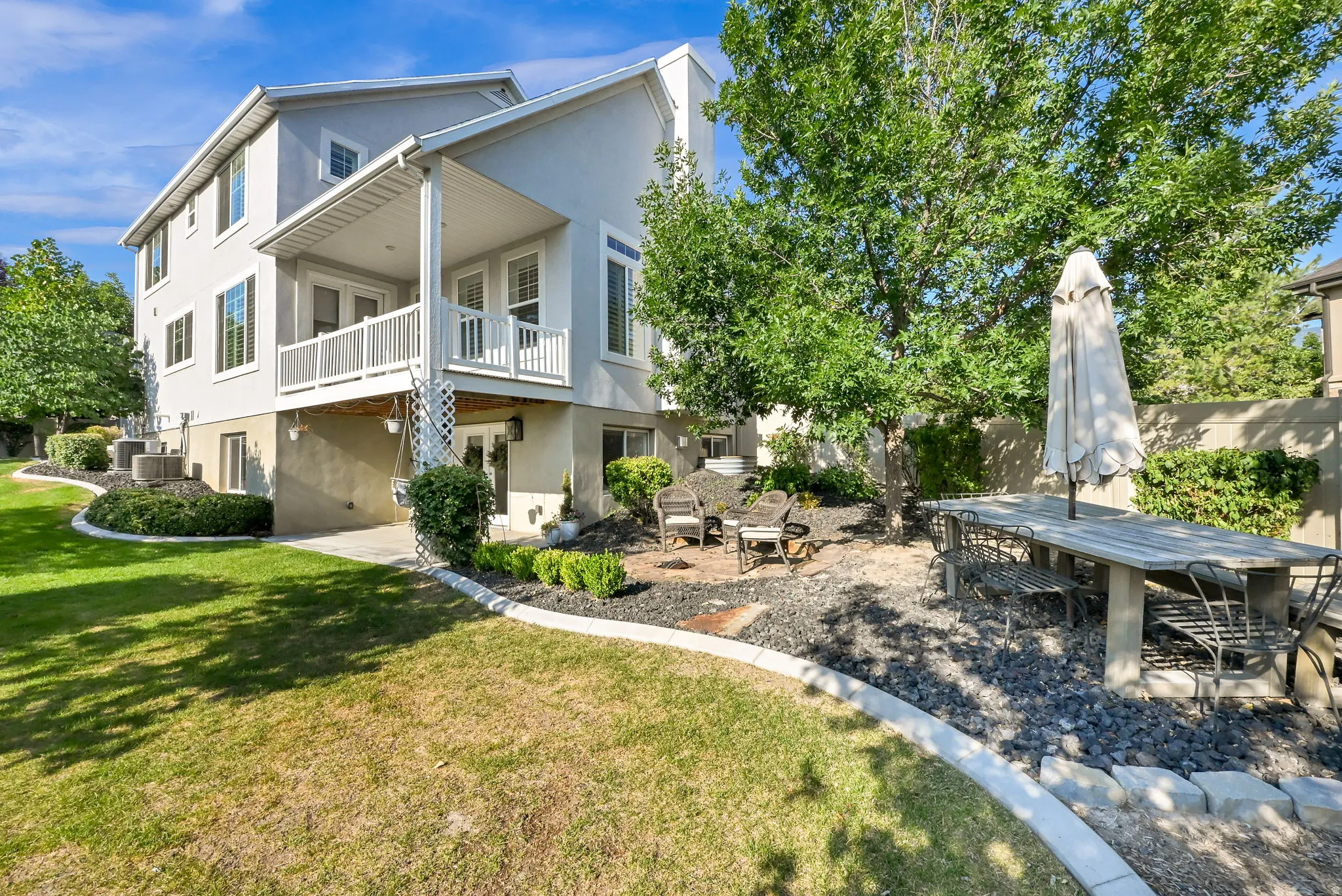 Rear exterior featuring a two-story stucco facade, second-story balcony with white railing, lower-level patio access, and landscaped yard with mature trees