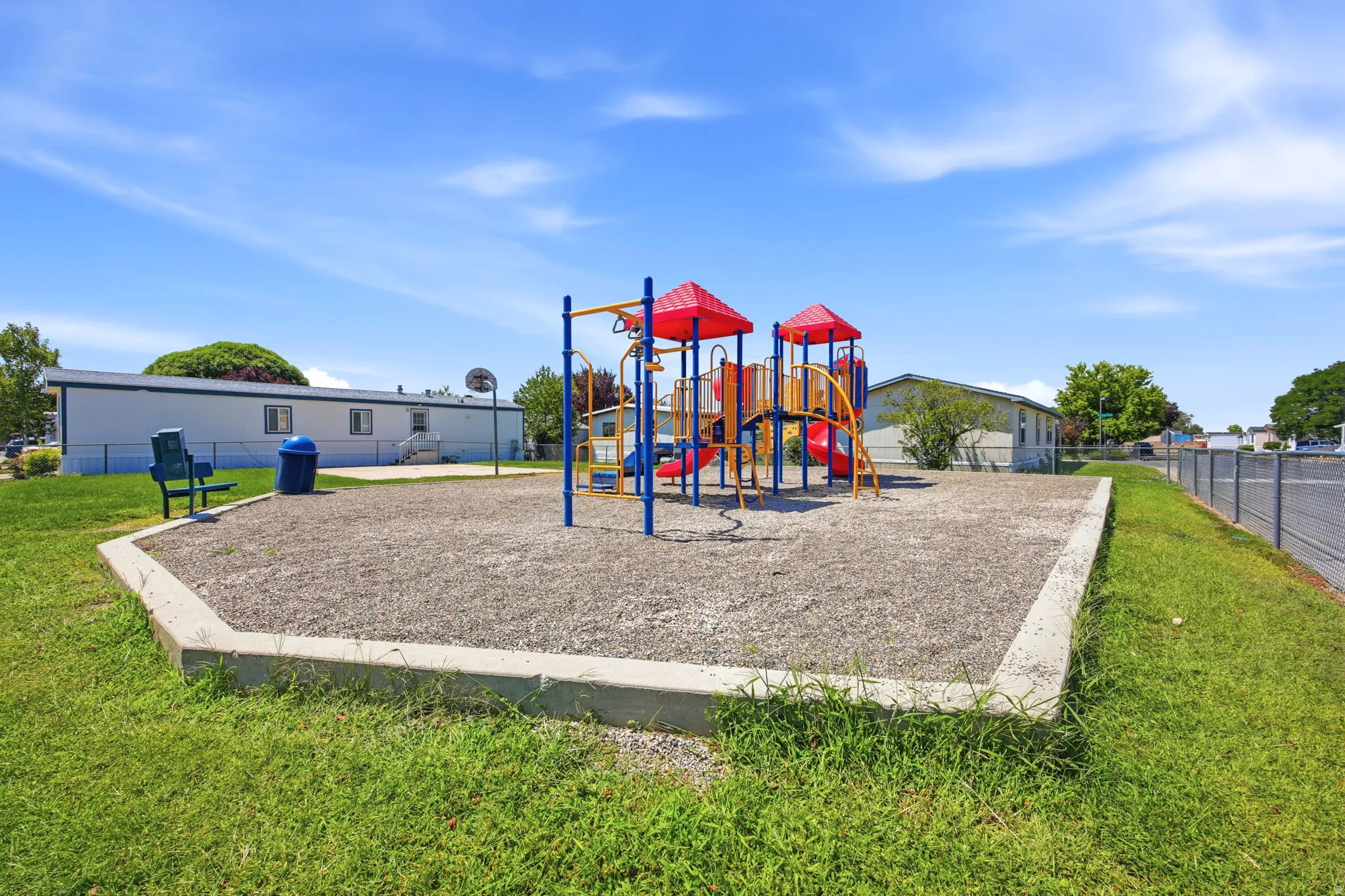Modern recreational area featuring a multi-level composite playground structure with red accents and blue support posts, set within a concrete-bordered safety surface