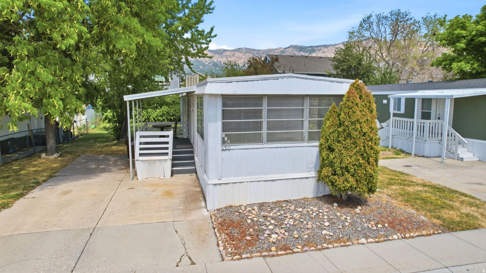 White mobile home with covered porch, concrete driveway, and established front landscaping