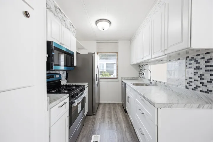 Galley kitchen featuring white cabinetry, wood-finish flooring, light-toned countertops, a mosaic tile backsplash, and a window providing natural light