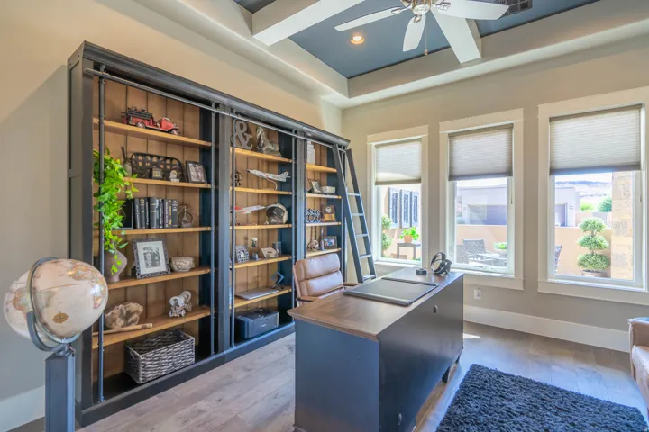Integrated shelving unit with rolling ladder, three large windows, a tray ceiling with a ceiling fan, and wood-finish flooring