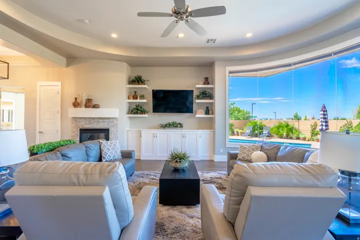 Spacious living area featuring a stone-clad fireplace, built-in shelving, a ceiling fan, and large curved windows overlooking a backyard pool