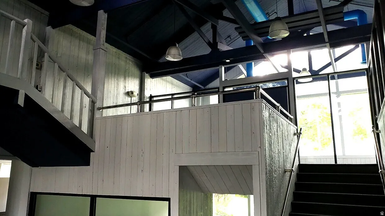 Spacious interior featuring a multi-level design with exposed ceiling beams, industrial pendant lighting, large windows, and a combination of white vertical paneling and dark-toned stairs with metal handrails