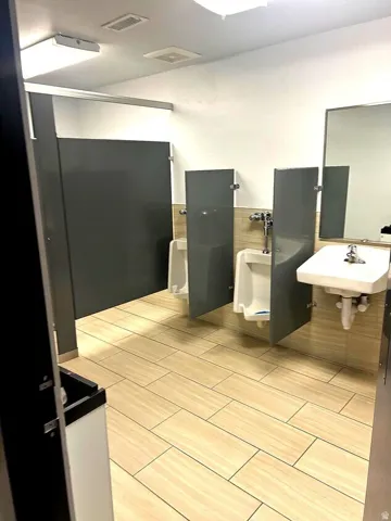 Commercial restroom featuring light-toned plank-style floor tiling, white walls, and a light wood-finish wainscoting