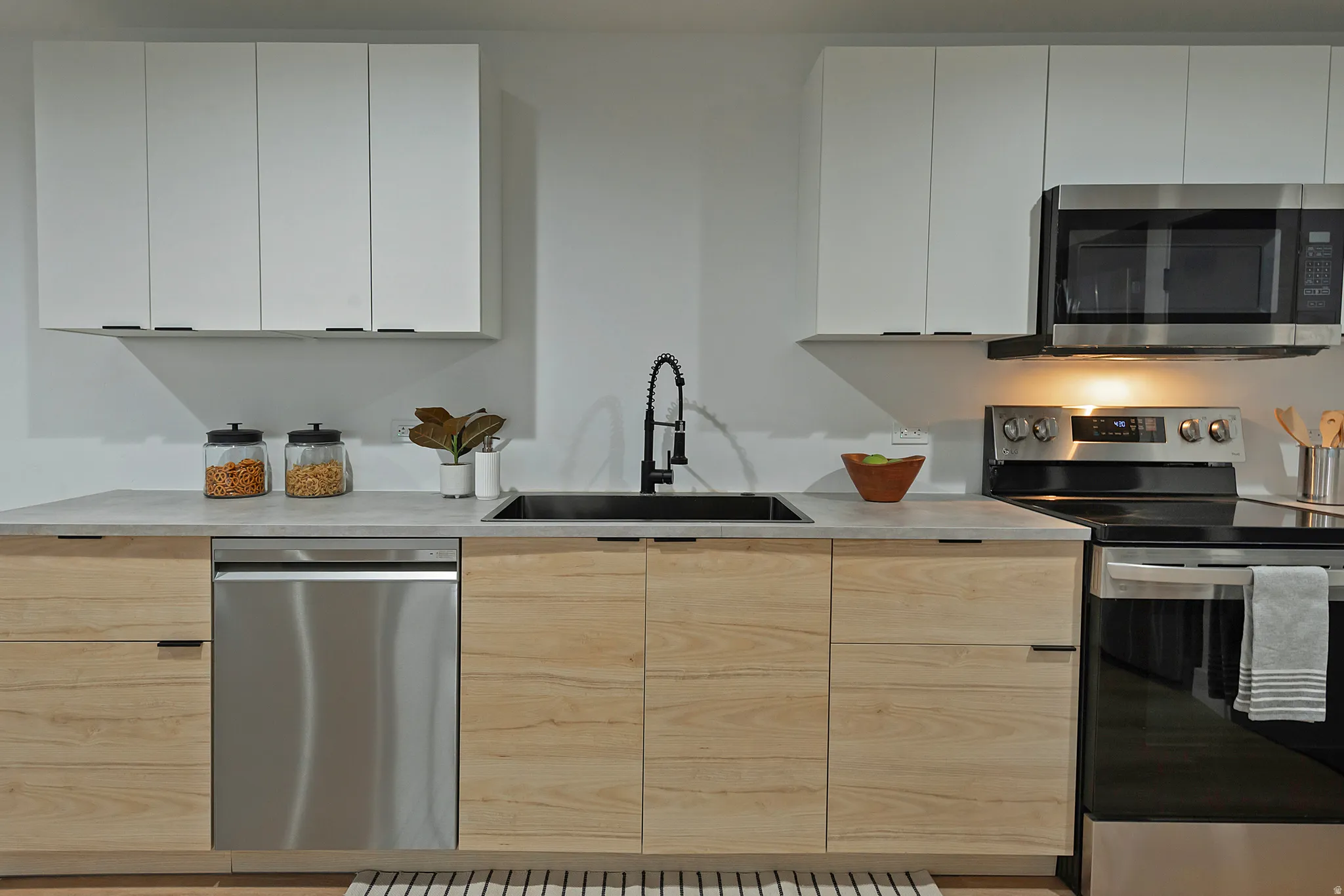 Kitchen featuring two-tone cabinetry with light wood-finish lower cabinets and white upper cabinets, a black sink with a gooseneck faucet, light-toned countertops, and stainless steel appliances including a dishwasher, range, and microwave
