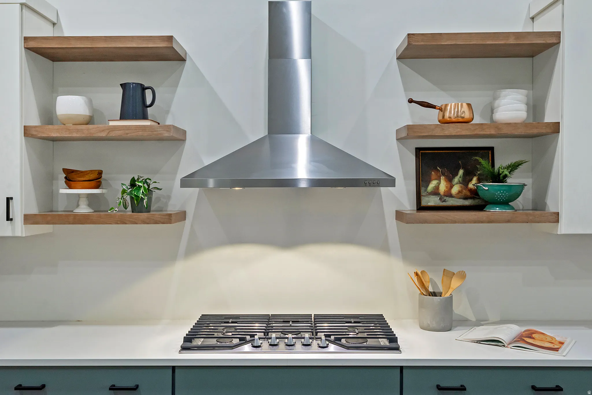 Gourmet kitchen featuring a stainless steel gas cooktop, professional-grade range hood, white countertops, teal base cabinetry with matte black hardware, and open wood-finish shelving