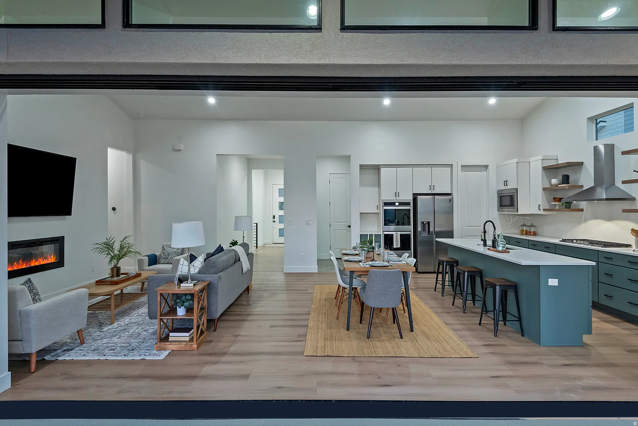 Expansive open-concept living area featuring wood-finish flooring throughout, a modern electric fireplace, a kitchen island with seating, a stainless steel range hood, and light-toned cabinetry