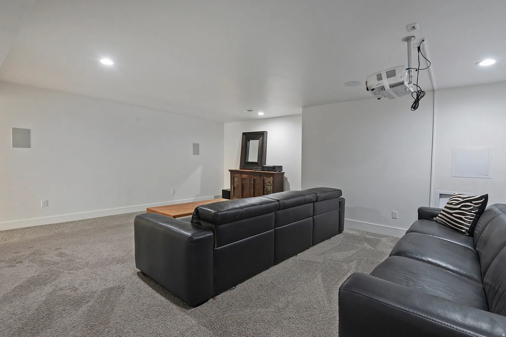 Spacious media room featuring recessed lighting, a ceiling-mounted projector, wall-mounted speakers, and light gray carpet flooring