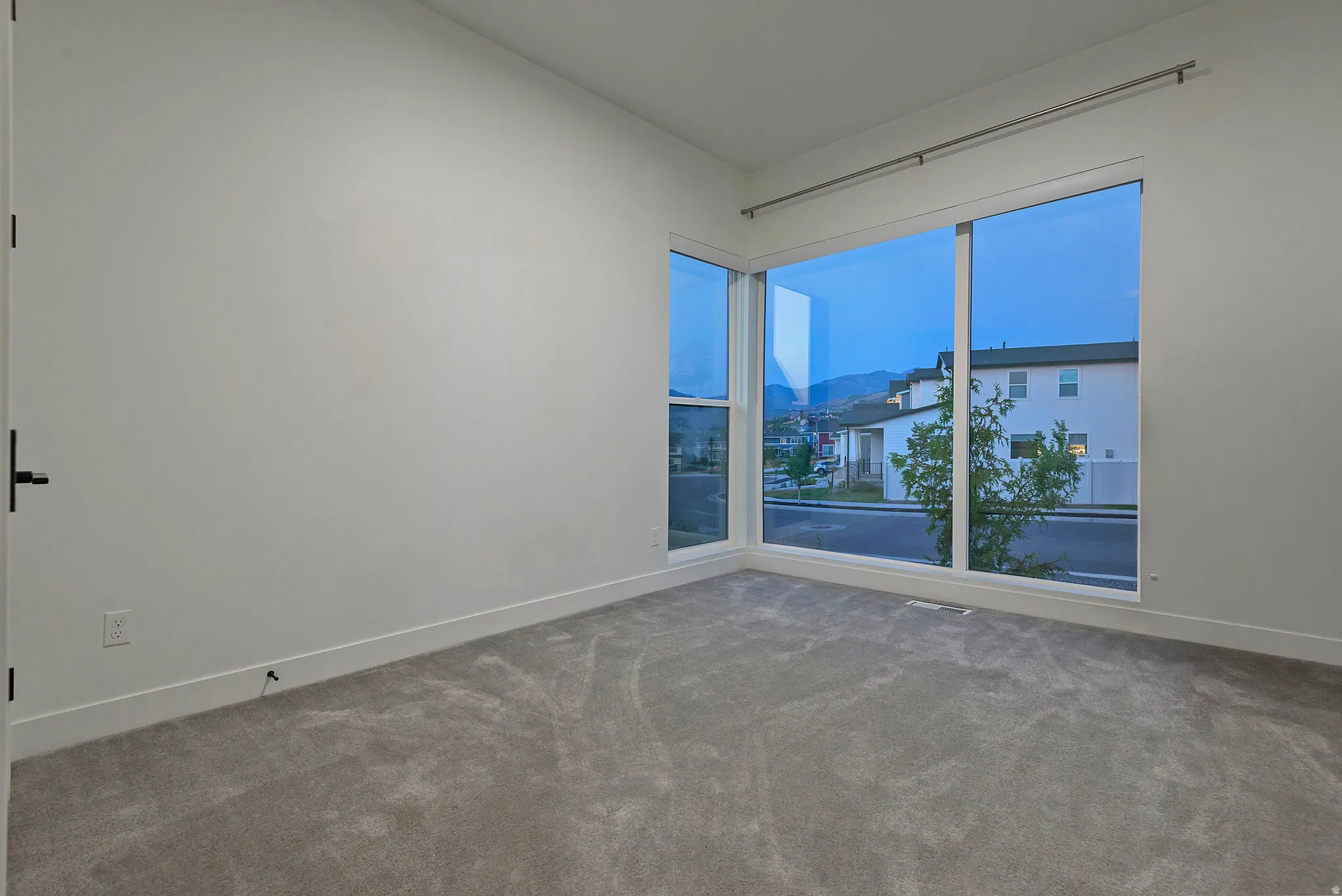 Spacious interior room featuring large corner windows, neutral wall tones, and gray carpeting