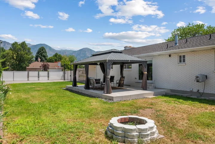 Spacious backyard featuring a covered patio with a dark-frame gazebo, a paver fire pit area, a white privacy fence, and a mountain backdrop