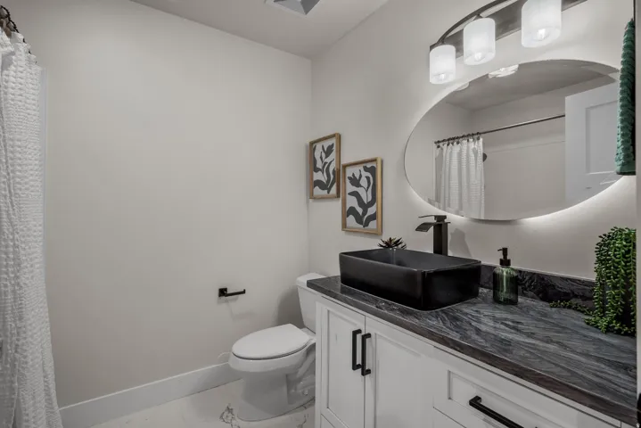 Bathroom featuring a rectangular vessel sink, dark stone countertop, white shaker-style cabinetry, and an illuminated oval mirror