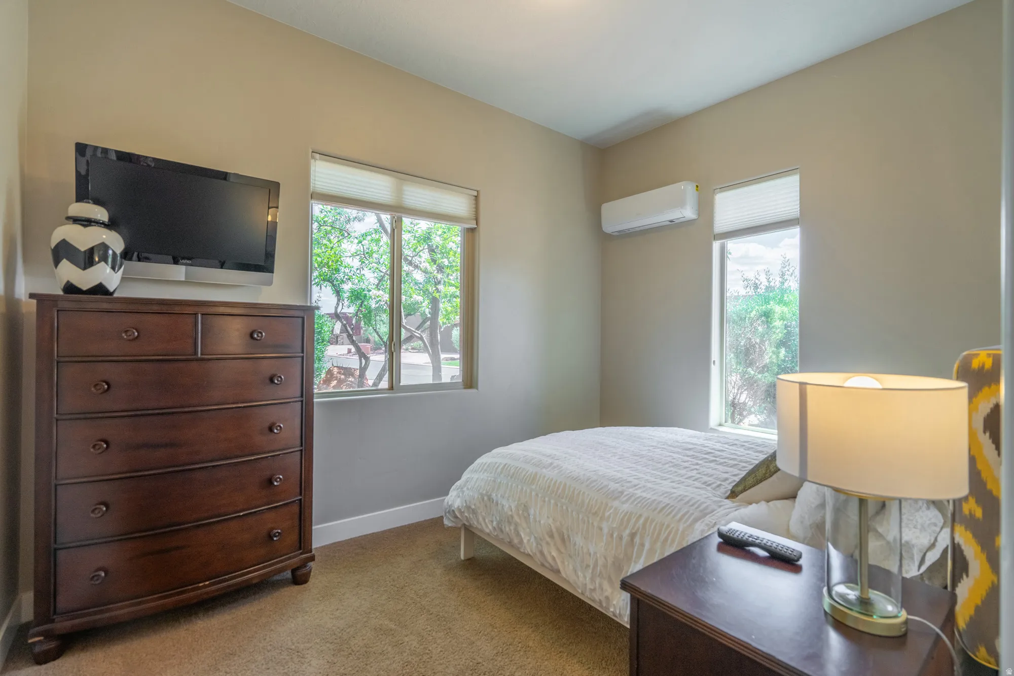 Carpeted room with neutral-toned walls, featuring a window with privacy blinds, a wall-mounted climate control unit, and a built-in wall-mounted television