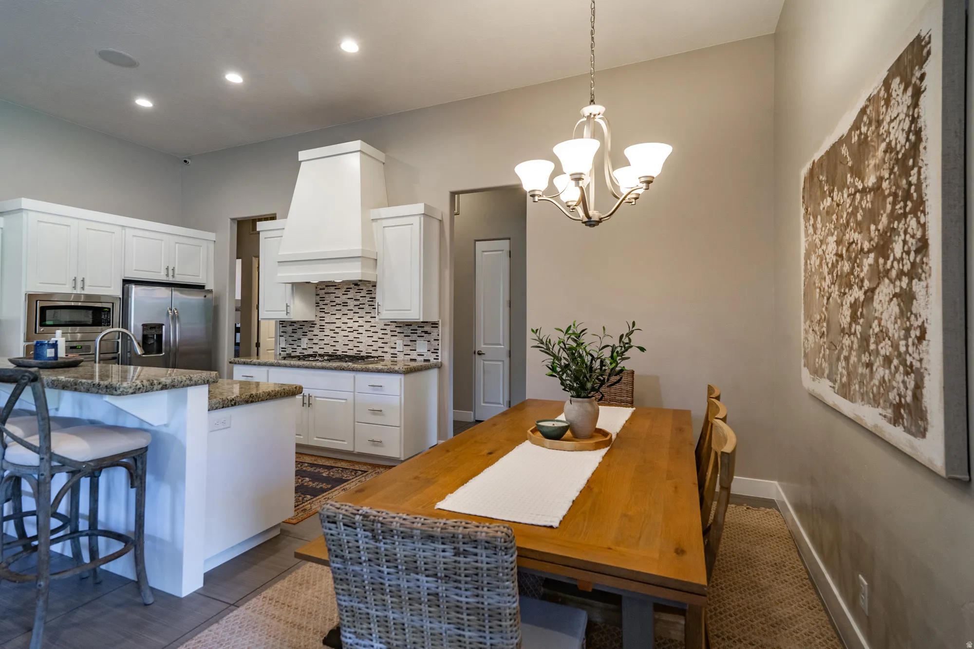 Open-concept kitchen and dining area featuring white cabinetry, granite countertops, a patterned tile backsplash, stainless steel appliances, and a central chandelier