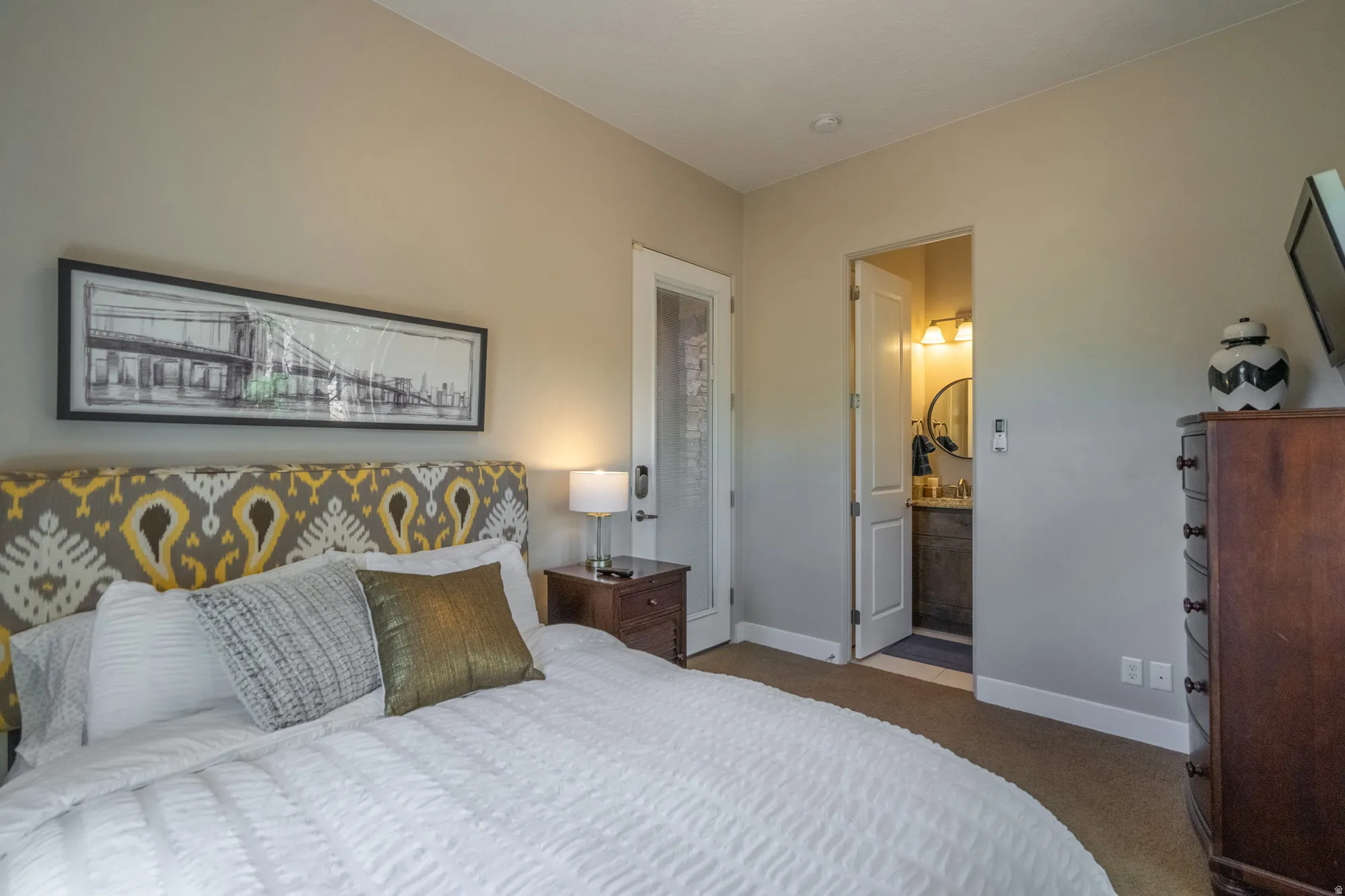 Neutral-toned bedroom featuring carpeted flooring and an adjacent bathroom with a vanity and circular mirror