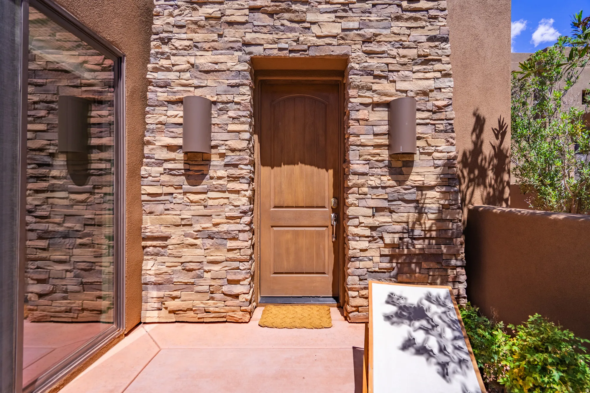 Entryway featuring stacked stone facade, natural wood door, two cylindrical sconces, stucco wall finish, and established landscaping