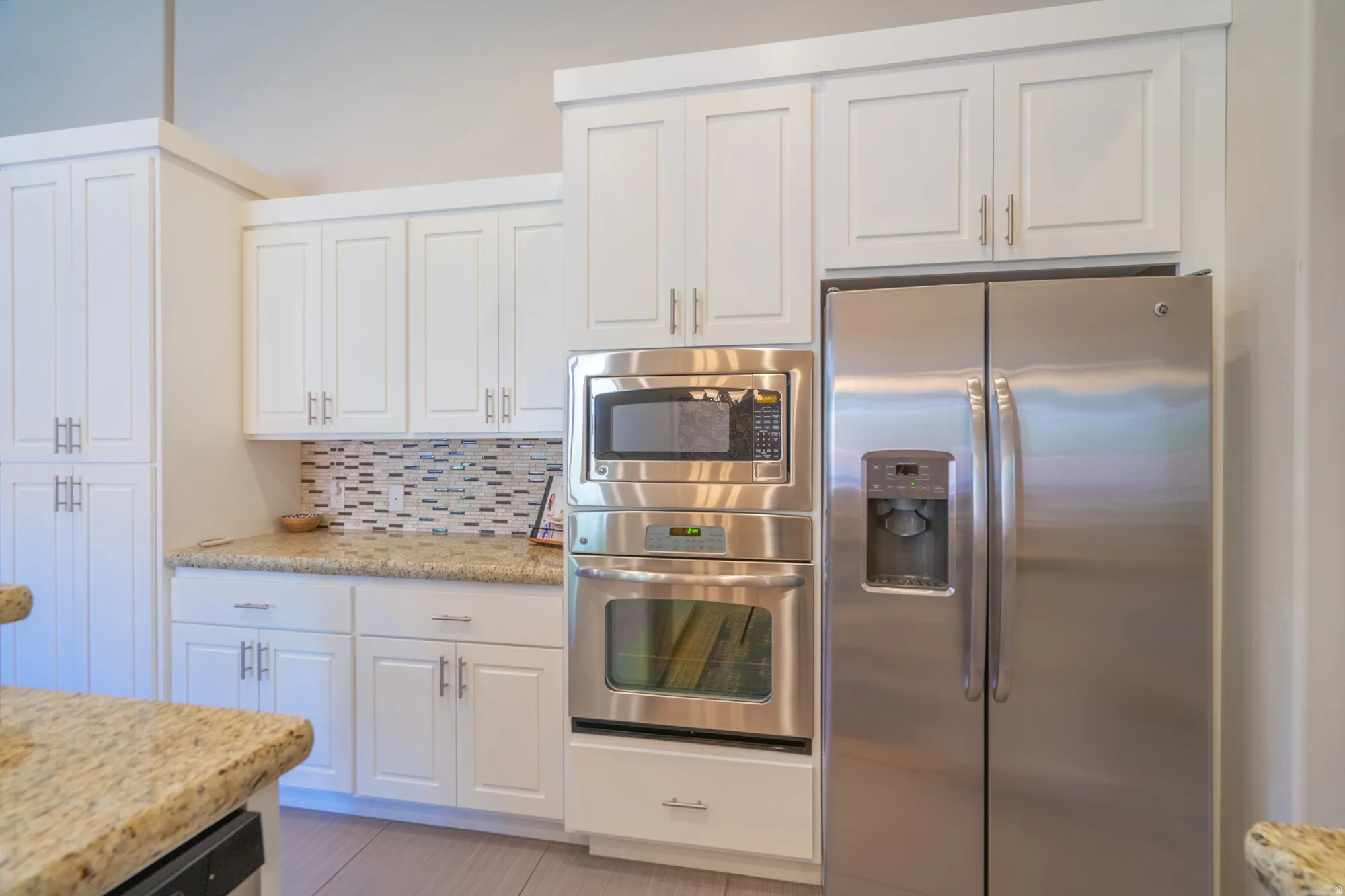 Kitchen featuring white shaker-style cabinetry with brushed nickel hardware, light-toned granite countertops, and a mosaic tile backsplash