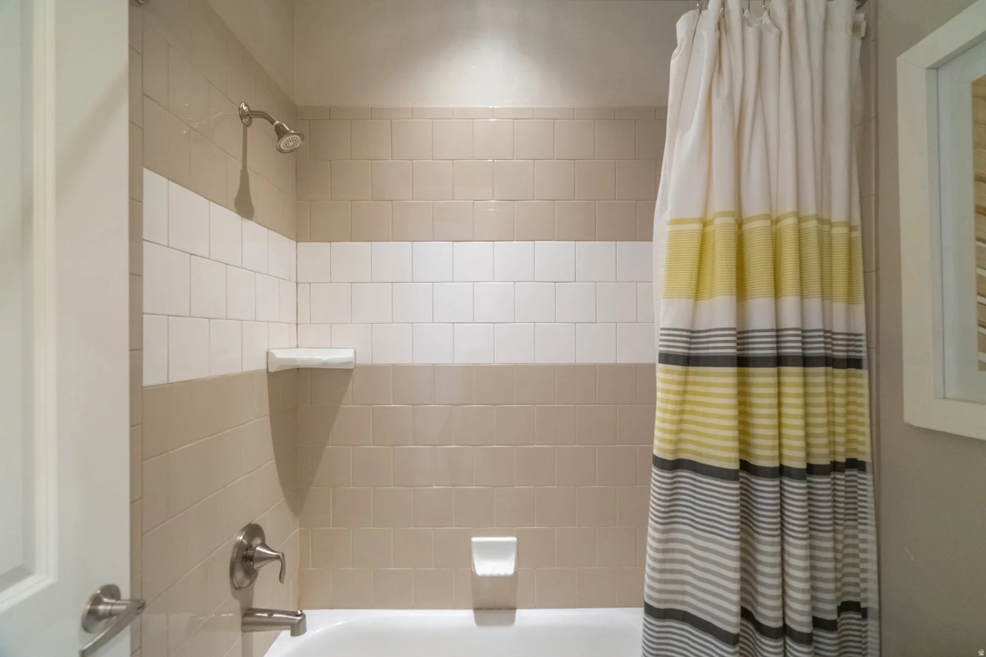 Bathtub and shower combination featuring neutral-toned tiled walls with a white accent band, brushed nickel fixtures, and a wall-mounted shower head