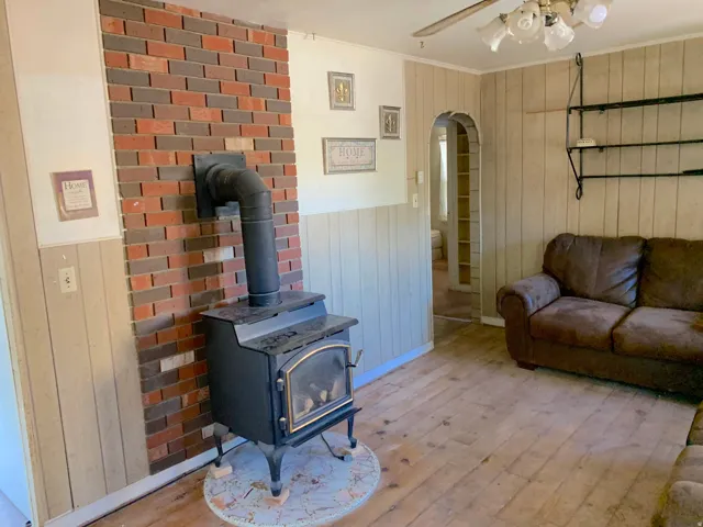 Wood-burning stove with brick surround, wood-finish flooring, paneled walls, ceiling fan, and arched doorway