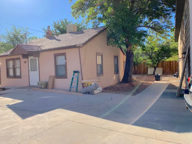Single-story stucco exterior with a gabled roof, prominent mature tree, and concrete driveway leading to a rear yard