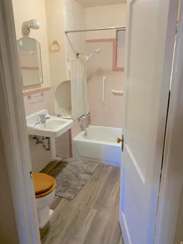 Bathroom featuring a white bathtub with a shower, a wall-mounted sink, and light wood-finish flooring