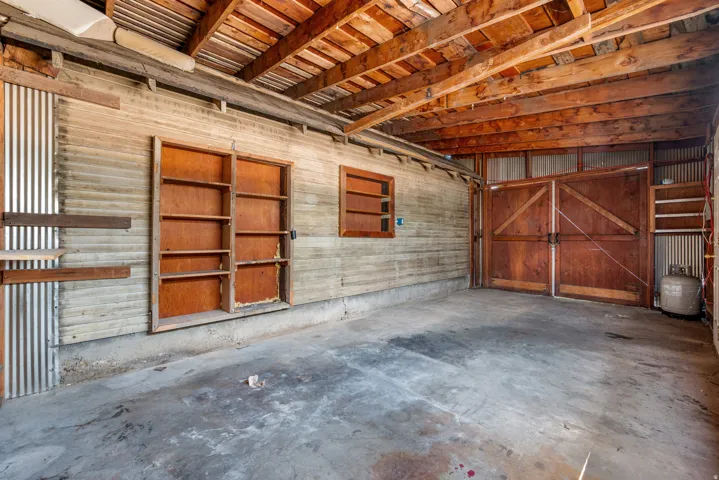 Spacious interior featuring exposed wood ceiling beams, concrete flooring, corrugated metal siding, built-in shelving, and large wood double doors