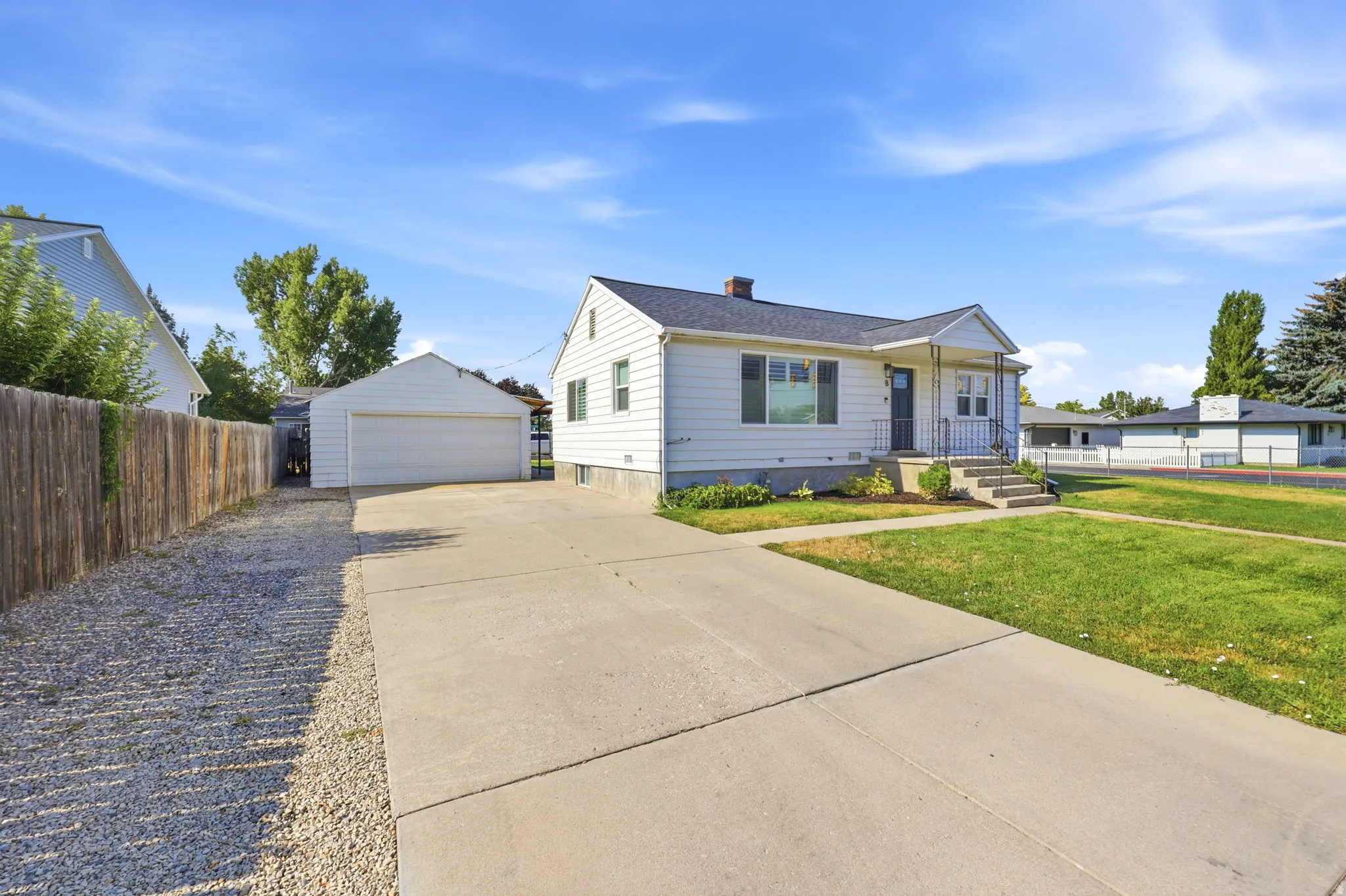 White siding residence featuring a covered front porch with iron railings, a concrete driveway, a detached garage, and a gravel side yard