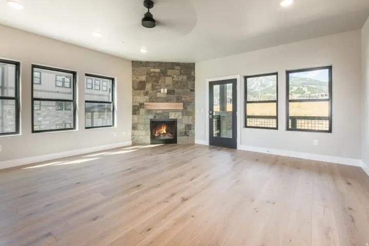 Open living area featuring light wood-finish flooring, a corner stone fireplace with a wood mantle, black-framed windows, and a black-framed glass door